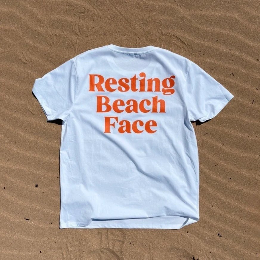 A white T-shirt laid flat on rippled sand, with RESTING BEACH FACE printed across the back in big orange letters
