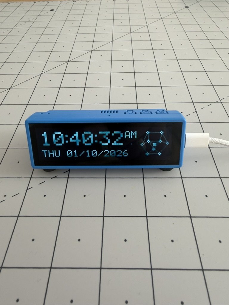 Tick on a cutting mat: a small blue case with a long, thin screen showing 10:40:32 AM in big blue dots, THU 01/10/2026 underneath and the cluster of linked nodes on the right, with a USB cable plugged into its side