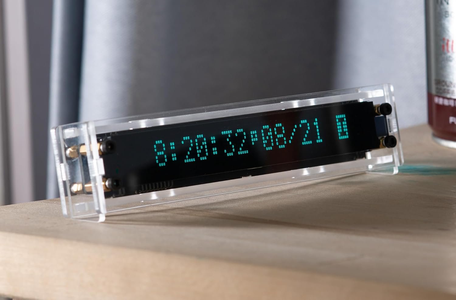 A real VFD clock in a clear acrylic case on a wooden shelf, showing the time and date in glowing blue-green dots, with the faint grid of unlit dots visible behind them